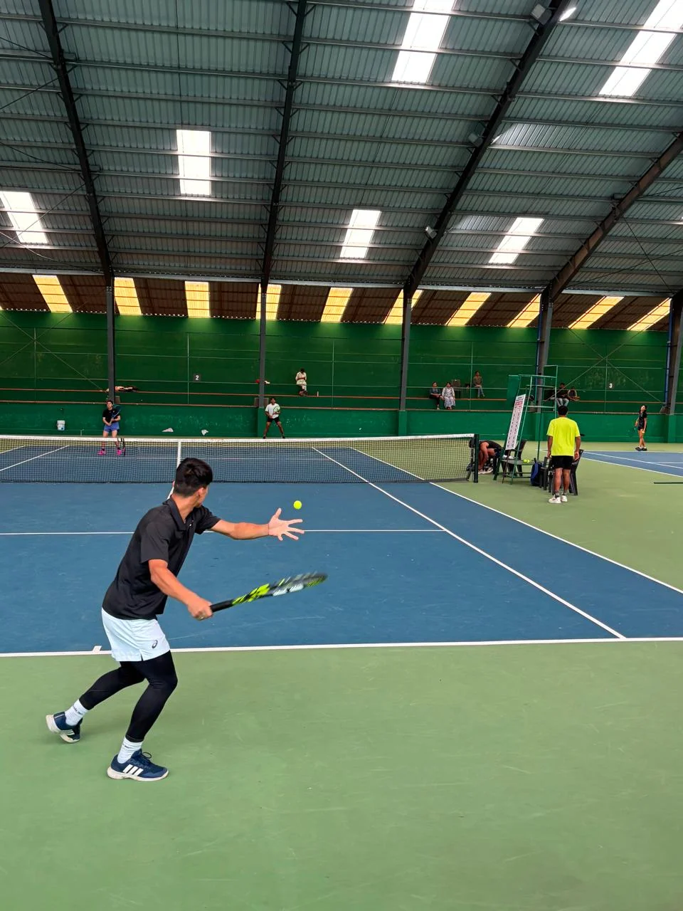 A player taking a forehand on an indoor hard court with matches running on the courts behind
